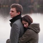 woman hugging man near body of water and trees during daytime