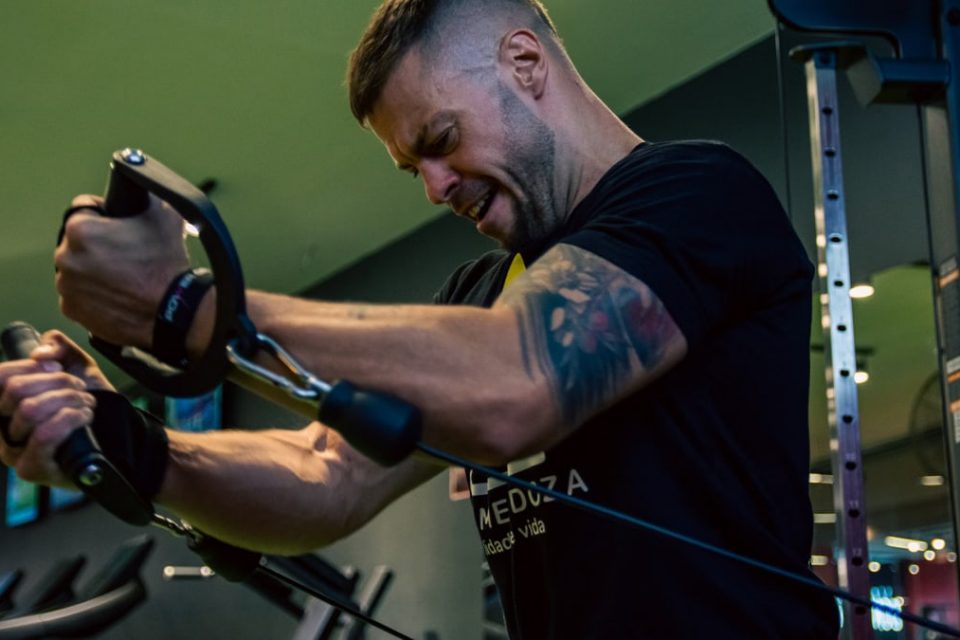 Muscular man with tattoos exercising on a cable machine in a gym, showcasing strength and determination.